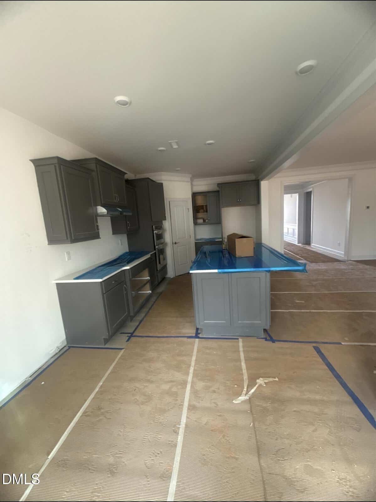 Modern kitchen with gray cabinets, blue protective film on countertops, and cardboard covering the floor. An island sits in the center, and adjacent rooms are visible through open doorways.