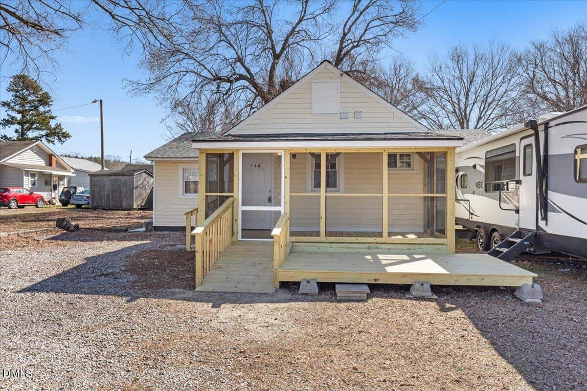 A small, single-story tan house with a gable roof, front porch with wooden railing, and steps. Gravel yard, bare trees, a shed to the left, parked vehicles, and clear blue sky are visible in the background.