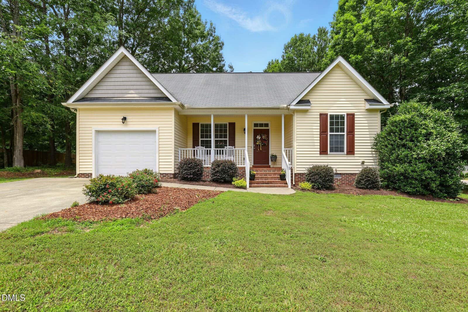 Single-story light yellow house with front porch, a white garage door on the left, brown shutters, steps leading to the main entrance, and landscaping with bushes and mulch; trees and grass surround the home at 75 Saddletree Drive in Franklinton.