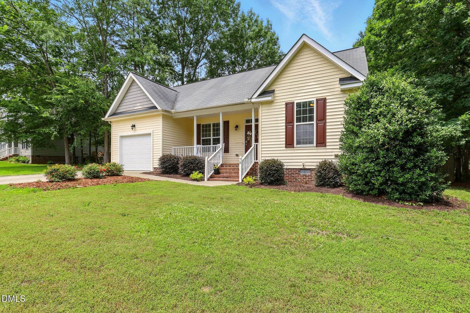 Single-story light yellow house with white trim, a front porch, brown shutters, and attached garage. The front yard has grass, bushes, mulch, and is surrounded by trees. The driveway is visible on the left side.