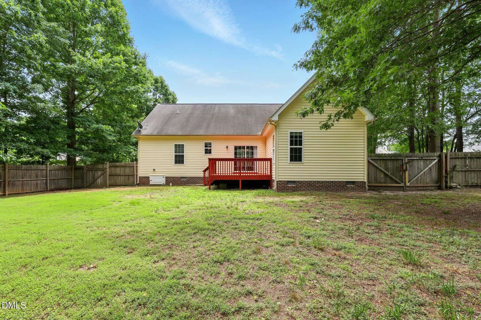 Single-story yellow house with white trim, red back porch, fenced yard, and a mix of green grass and bare patches. Tall trees surround the yard, and the sky is clear with wispy clouds.