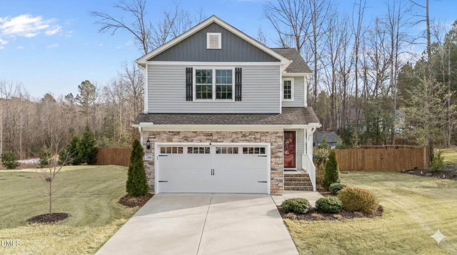 A two-story house with gray siding and stone accents, black shutters, and a white double garage sits on a manicured lawn bordered by shrubs and trees. A wooden fence runs along the right side of the backyard.