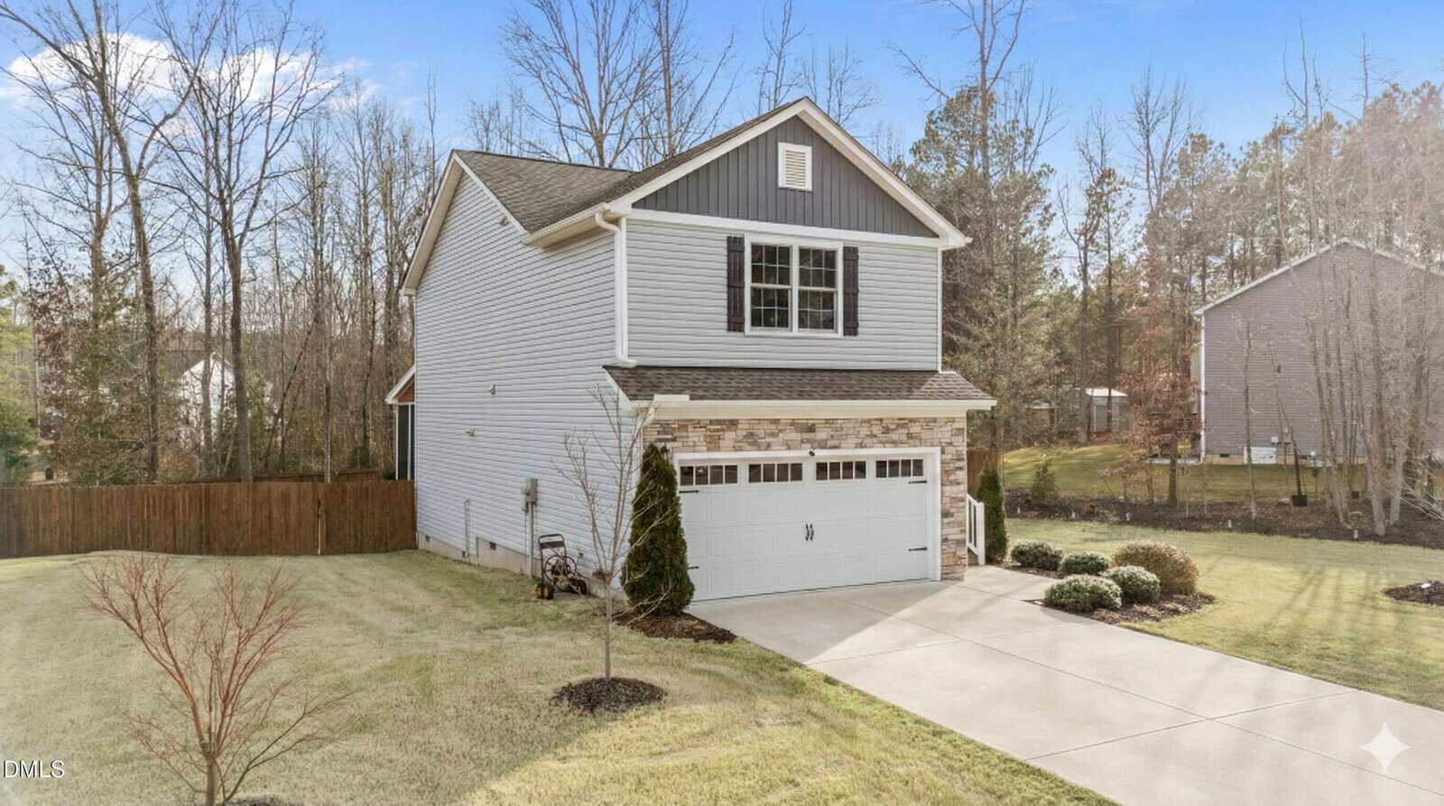 Two-story suburban house with gray siding, stone trim, white garage door, and a small front porch. Concrete driveway and trimmed lawn, with trees and a wooden fence in the backyard. Clear blue sky overhead.