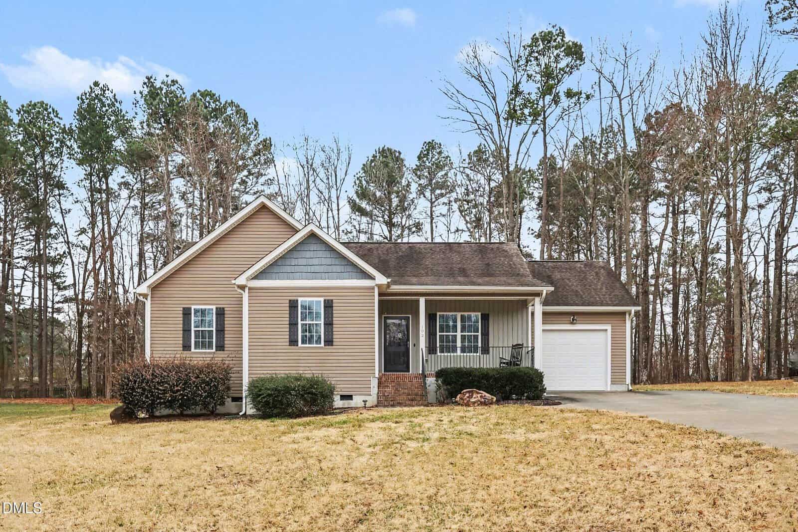 Single-story beige house with white trim, front porch, attached garage, and shingled roof. The house sits on a spacious, dry lawn bordered by tall trees, with a concrete driveway leading to the garage.