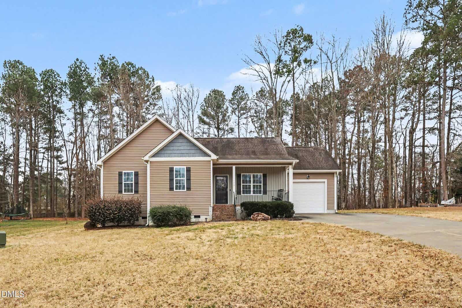 Single-story beige house with white trim, a front porch, attached one-car garage, and a concrete driveway. The yard is mostly brown grass, and the property is bordered by tall trees in the background under a blue sky.