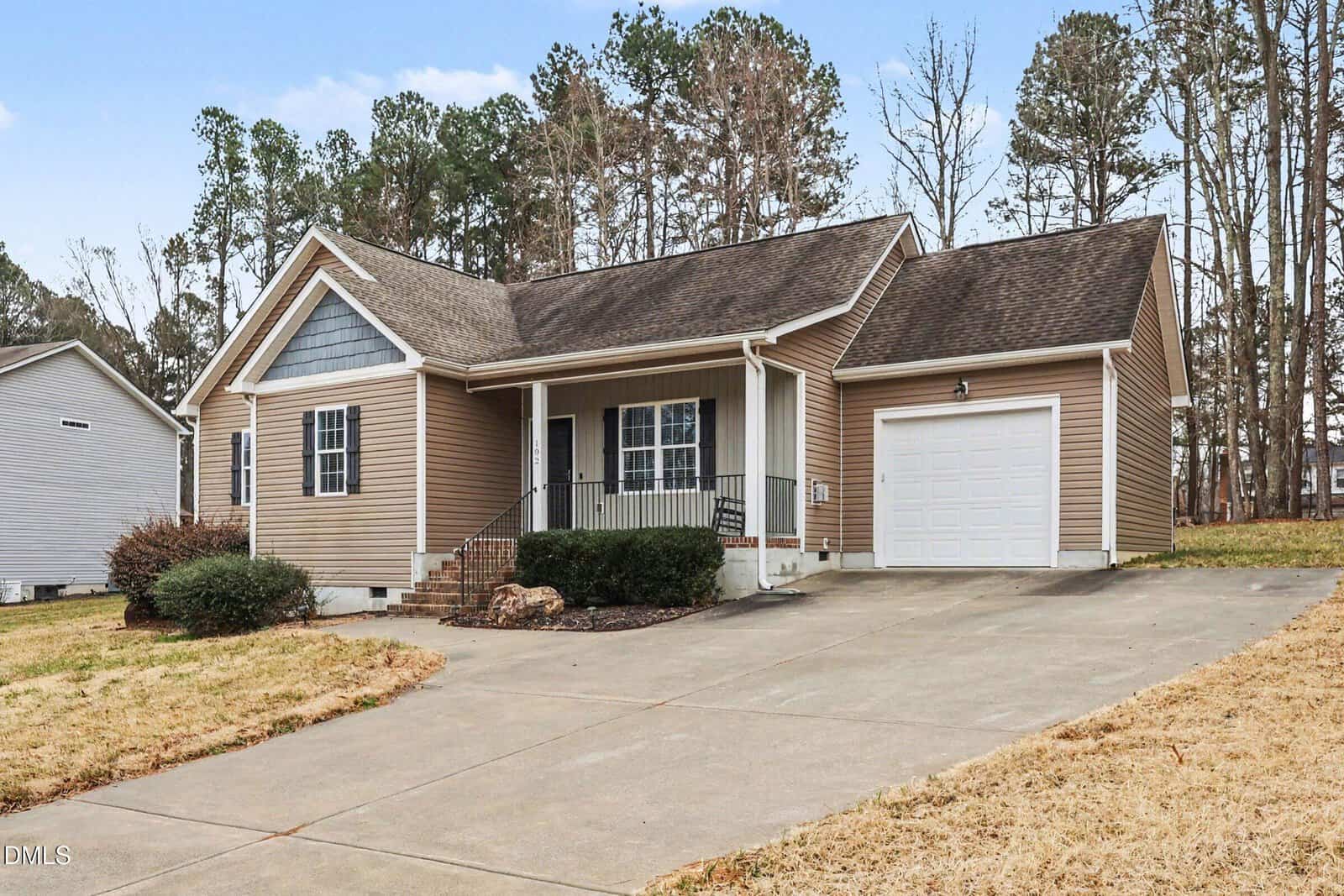 Single-story tan house with a front porch, attached garage, and sloped driveway. The yard is mostly brown grass with some bushes. Trees are in the background under a partly cloudy sky.