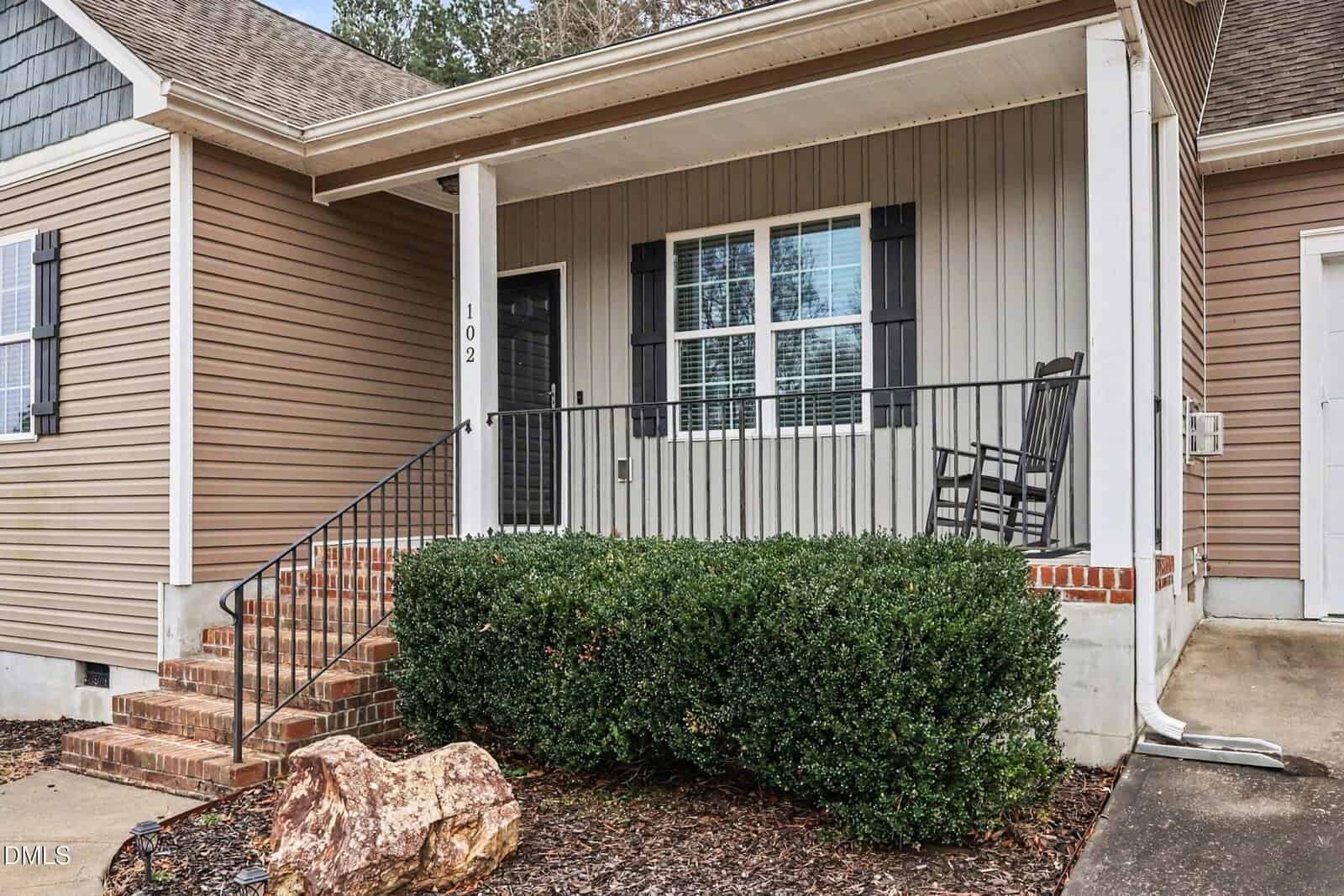 Front porch of a house with tan siding, black door, white trim, black shutters, a rocking chair, brick steps, metal railing, green bushes, and a large rock in the foreground.