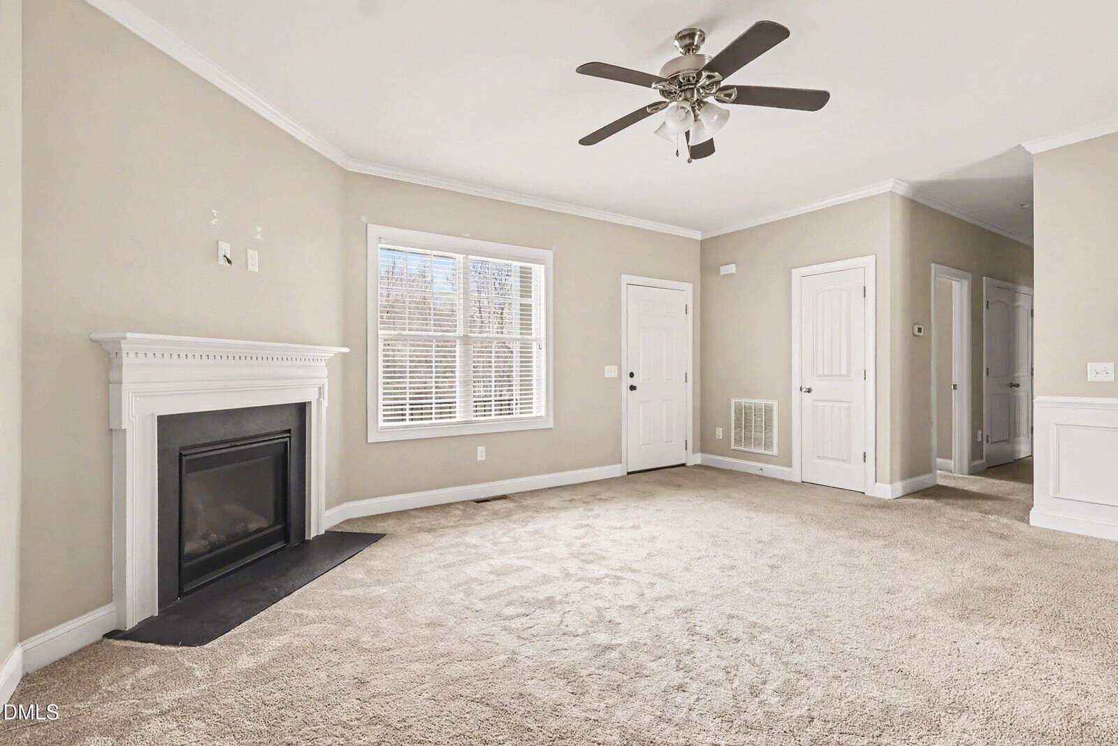 Bright living room with beige carpet, light taupe walls, a white fireplace, large window, white doors, ceiling fan, and crown molding. Room is empty and well-lit with natural light.