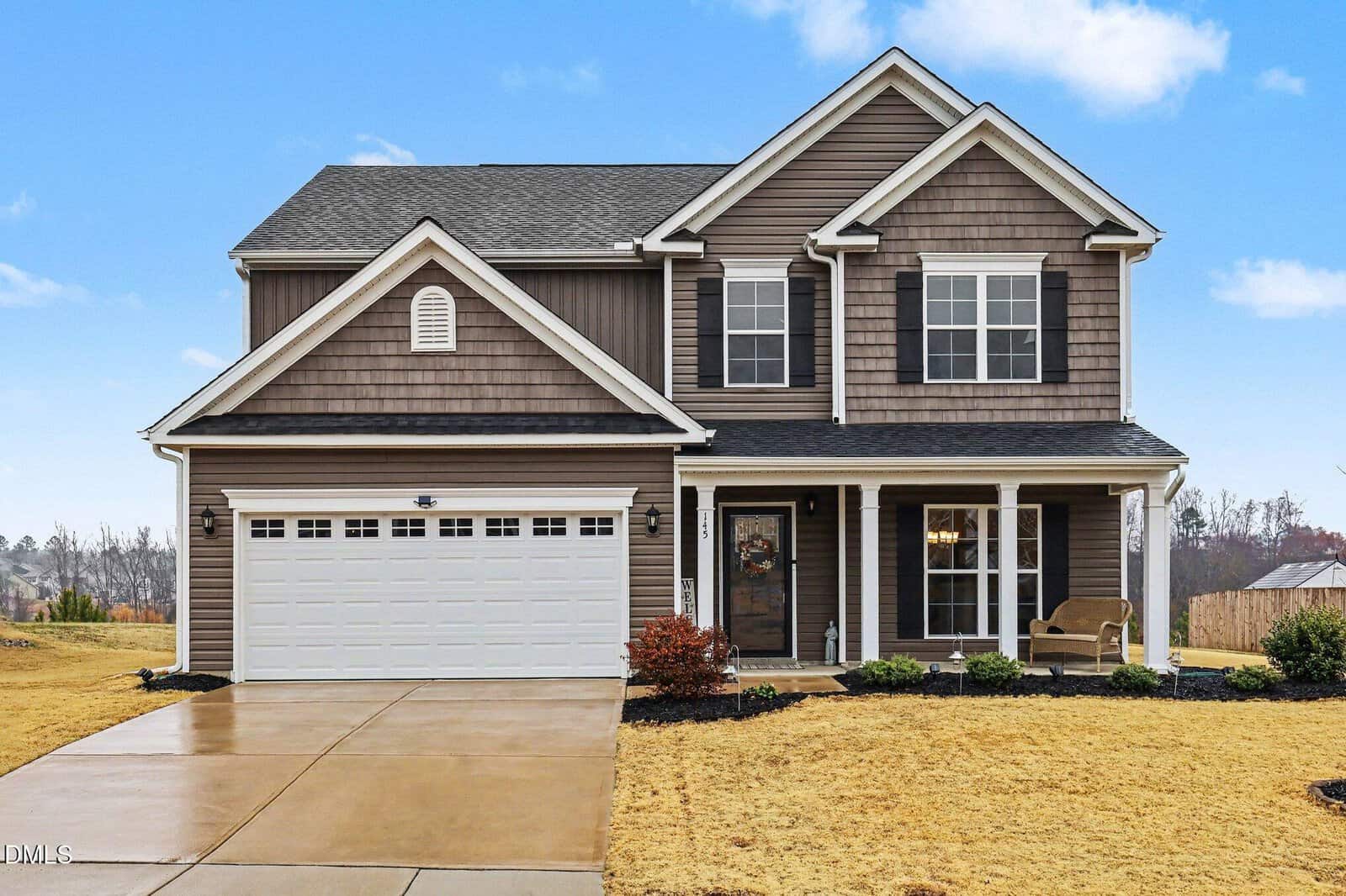 Two-story suburban house with brown siding, white trim, a covered front porch, black shutters, and a two-car garage. The driveway and lawn are in the foreground under a clear blue sky.