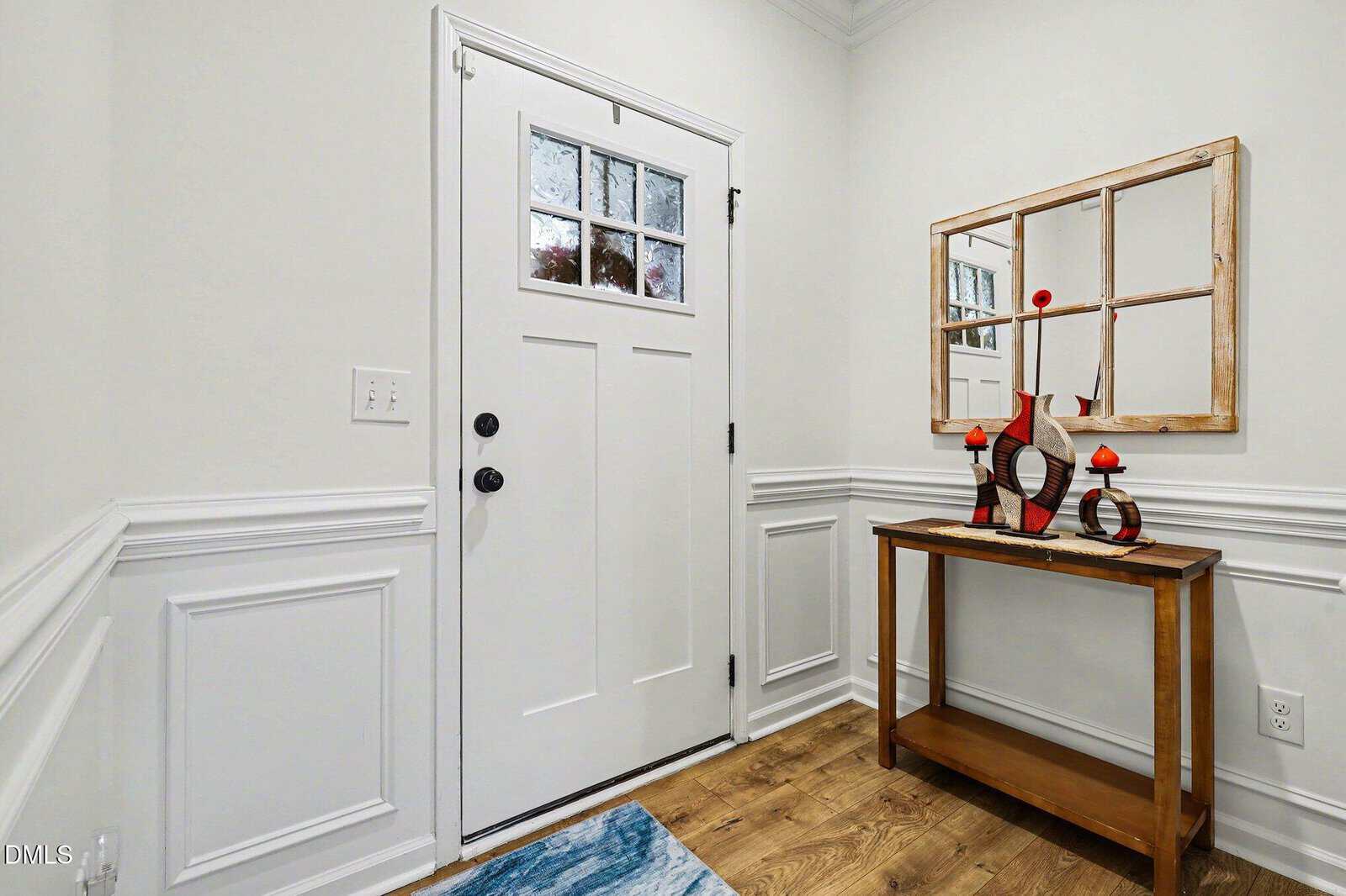 A small entryway with a white door, wood console table, decorative mirror, red candle holders, and abstract decor; white paneled walls and wood flooring are visible.