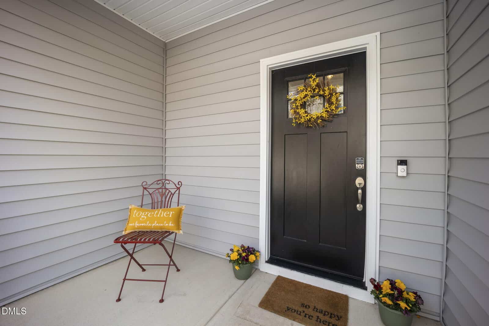 A black front door with a yellow wreath, a brown doormat reading “so happy you’re here,” a metal chair with a yellow pillow, and two flower pots on the porch of a house with gray siding.