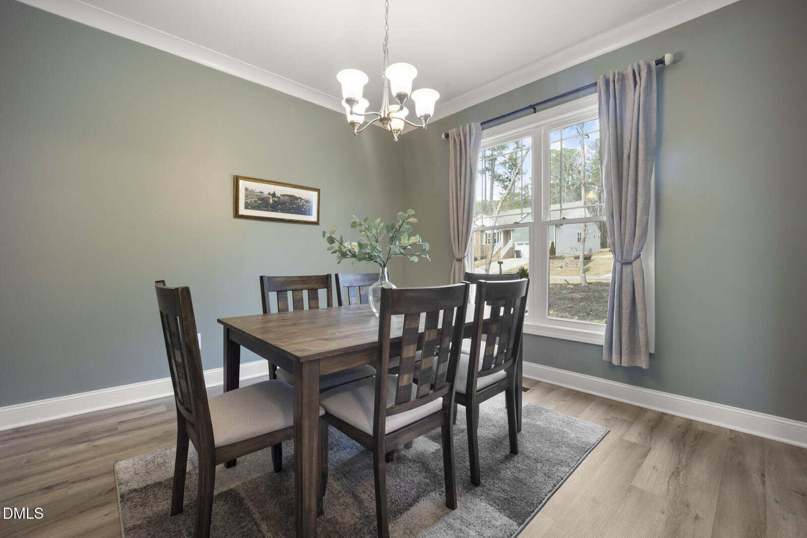 A dining room with a wooden table and six chairs, a small plant centerpiece, chandelier lighting, green walls, a framed picture, gray curtains, a large window, and wood flooring with a gray rug.