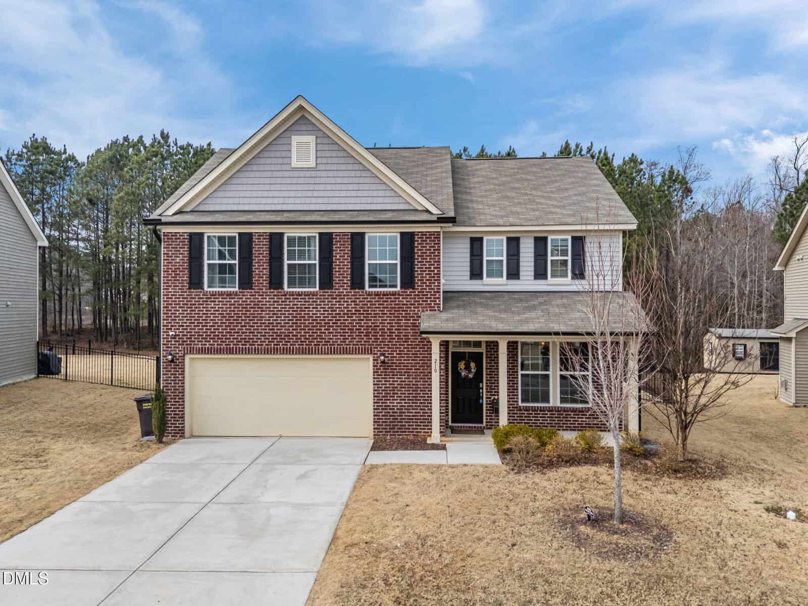 Two-story suburban house with red brick and beige siding exterior, black shutters, attached two-car garage, and a leafless tree in the front yard. The driveway and lawn are visible under a blue sky.