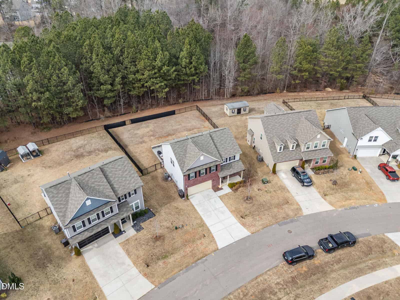 An aerial view of a suburban neighborhood showing five two-story houses with driveways, backyards, and cars parked along the curved street. A wooded area is visible behind the houses.