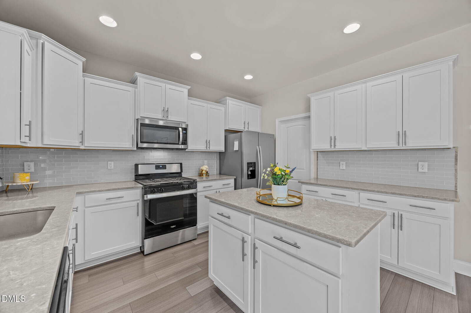 Modern kitchen with white cabinets, stainless steel appliances, light gray countertops, a central island with a decorative tray and potted plant, light wood flooring, and recessed ceiling lights.