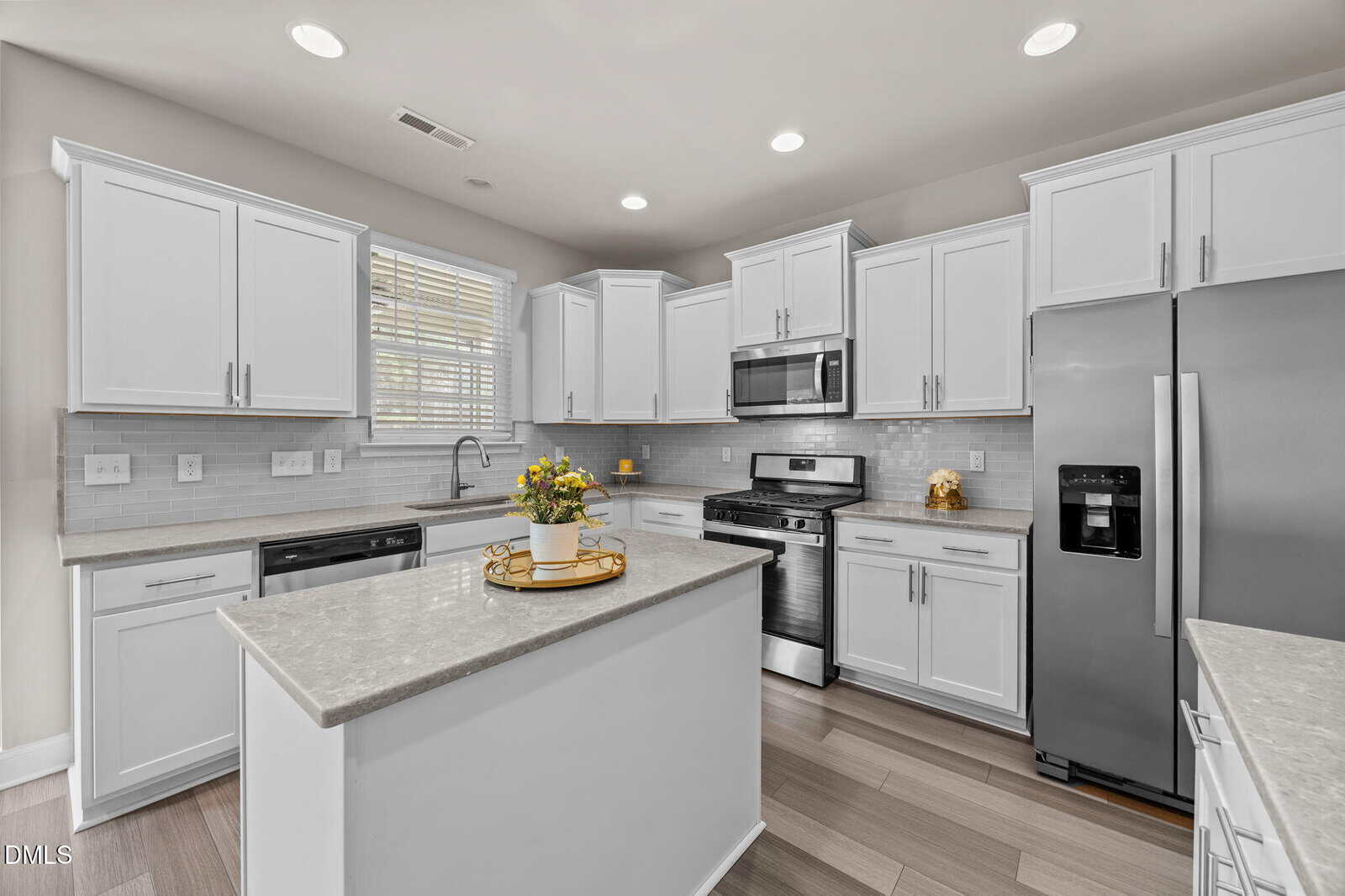 Modern kitchen with white cabinets, stainless steel appliances, a center island with a decorative tray and flowers, light gray countertops, tile backsplash, and wood-look flooring under recessed lighting.