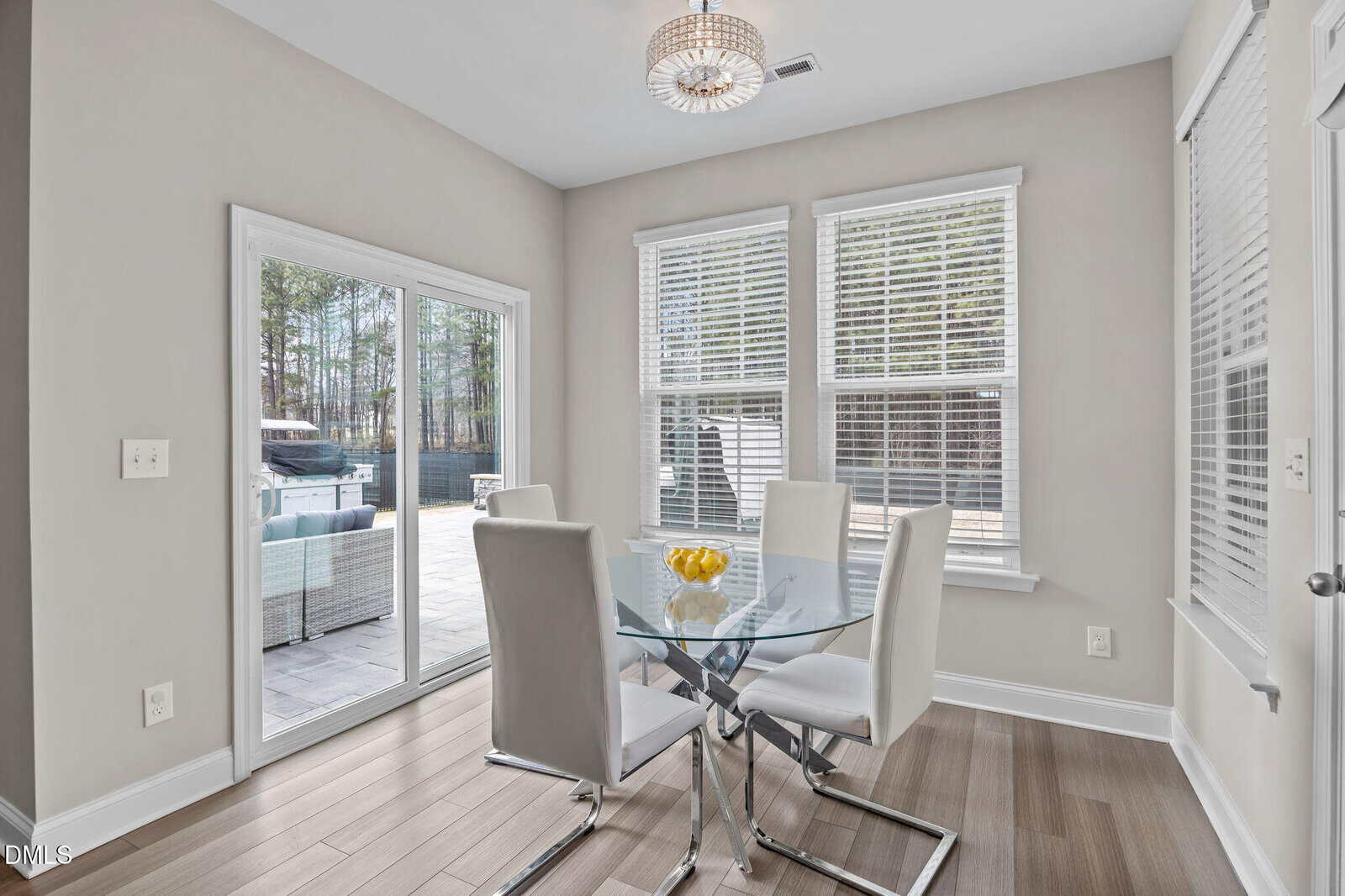 A dining area with a glass table and four white chairs, set on wood flooring. Windows and a sliding glass door provide views of an outdoor patio with seating and trees. A bowl of lemons sits on the table.