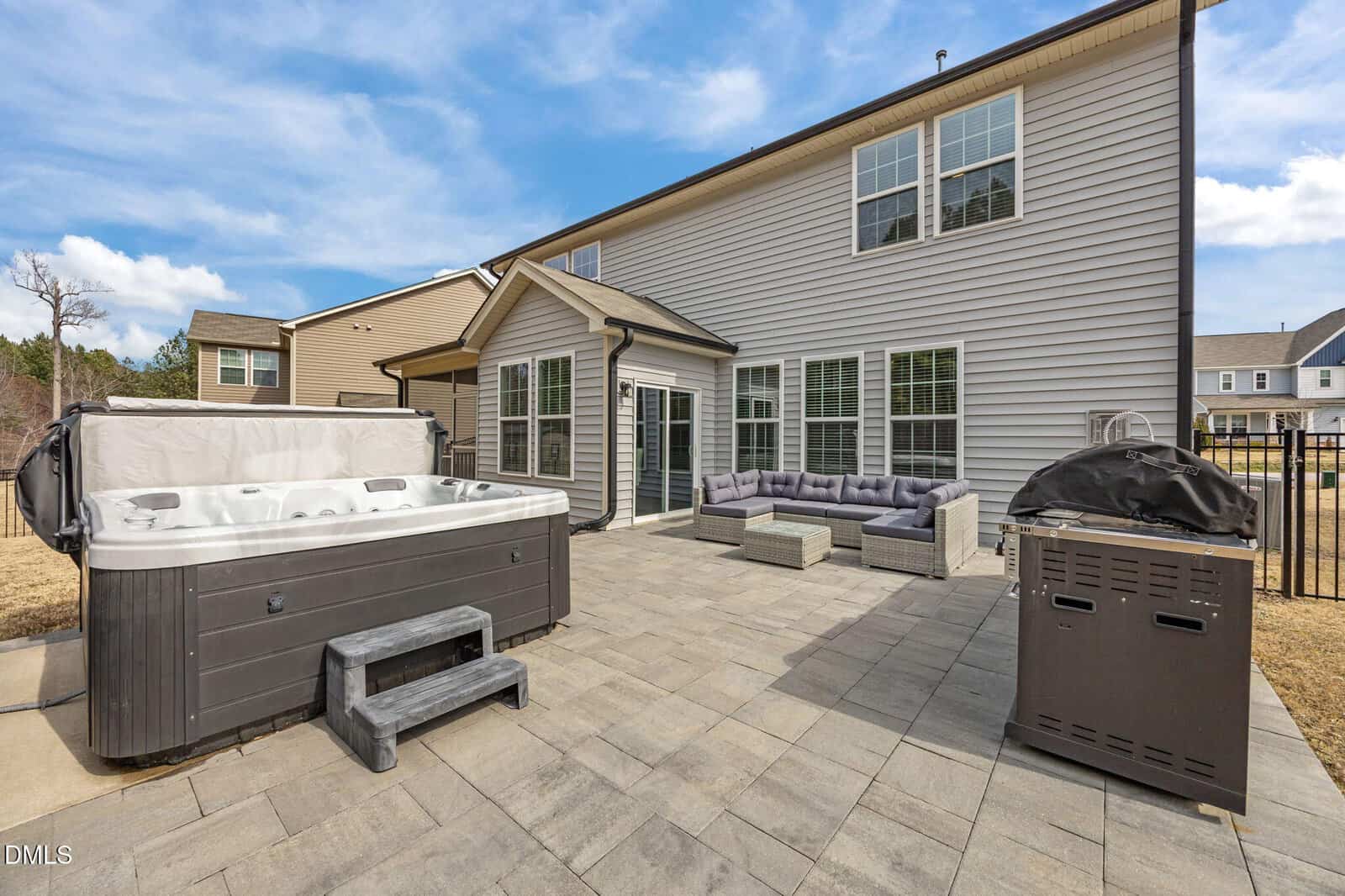 A backyard patio with a large hot tub, a gray outdoor sectional sofa, a grill, and stone pavers. The patio is attached to a two-story beige house with multiple windows.