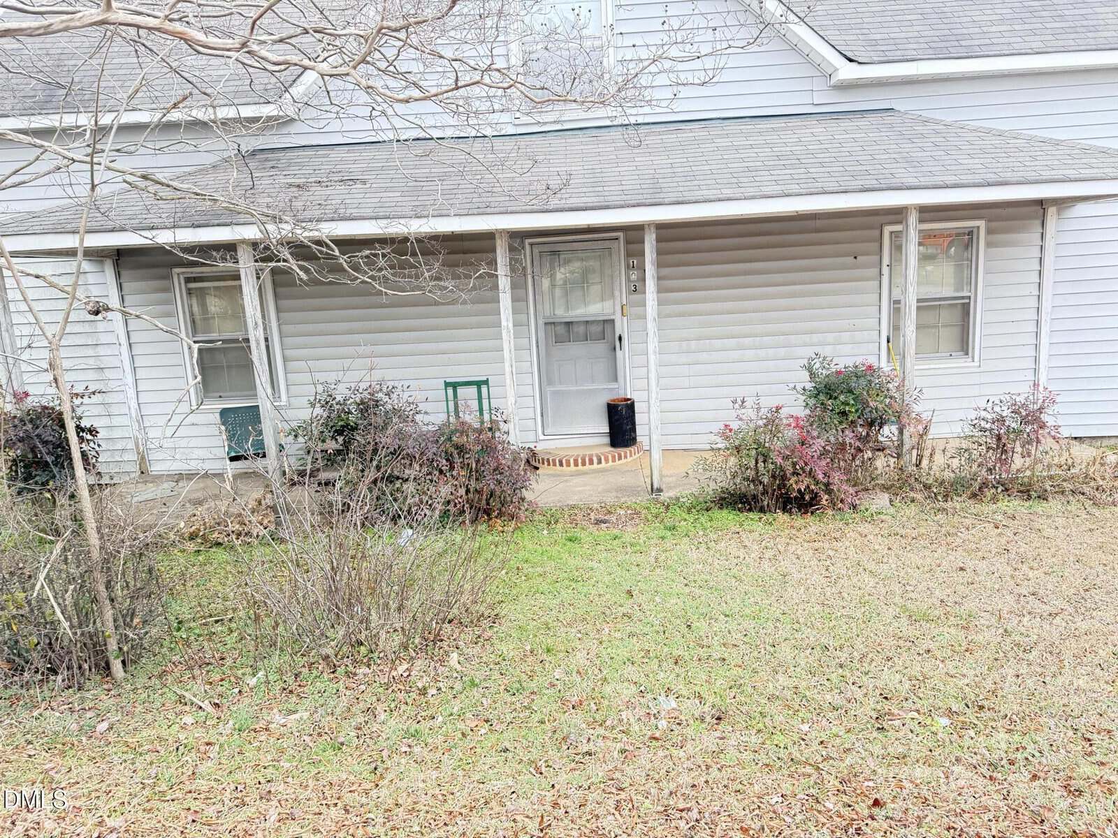 A one-story house with light gray siding, a covered front porch, two windows, a central door, and shrubs in the front yard. There is a small green bench and a black container near the door.