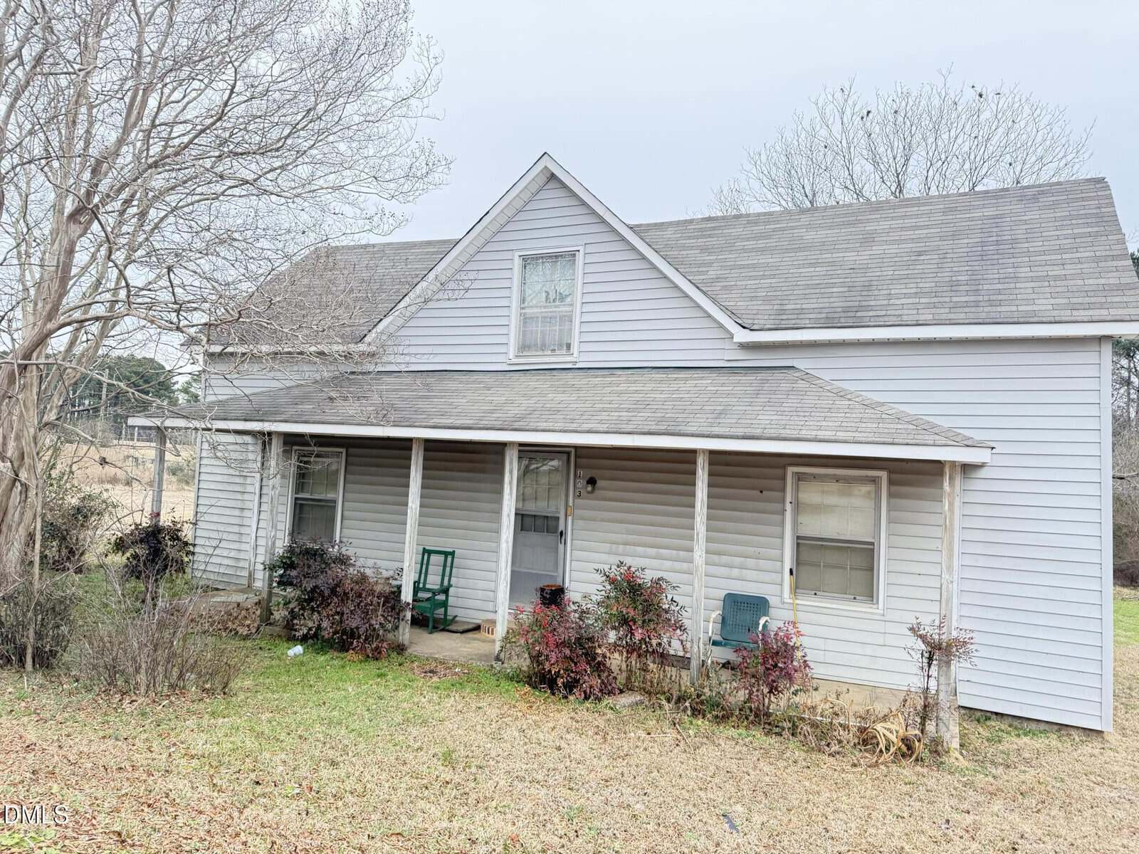A two-story light gray house with a covered front porch, a sloped roof, several windows, and a lawn with bushes and bare trees sits quietly on 103 Howard Harris Road in Franklinton. Two chairs and a green swing are on the porch. The sky is overcast.
