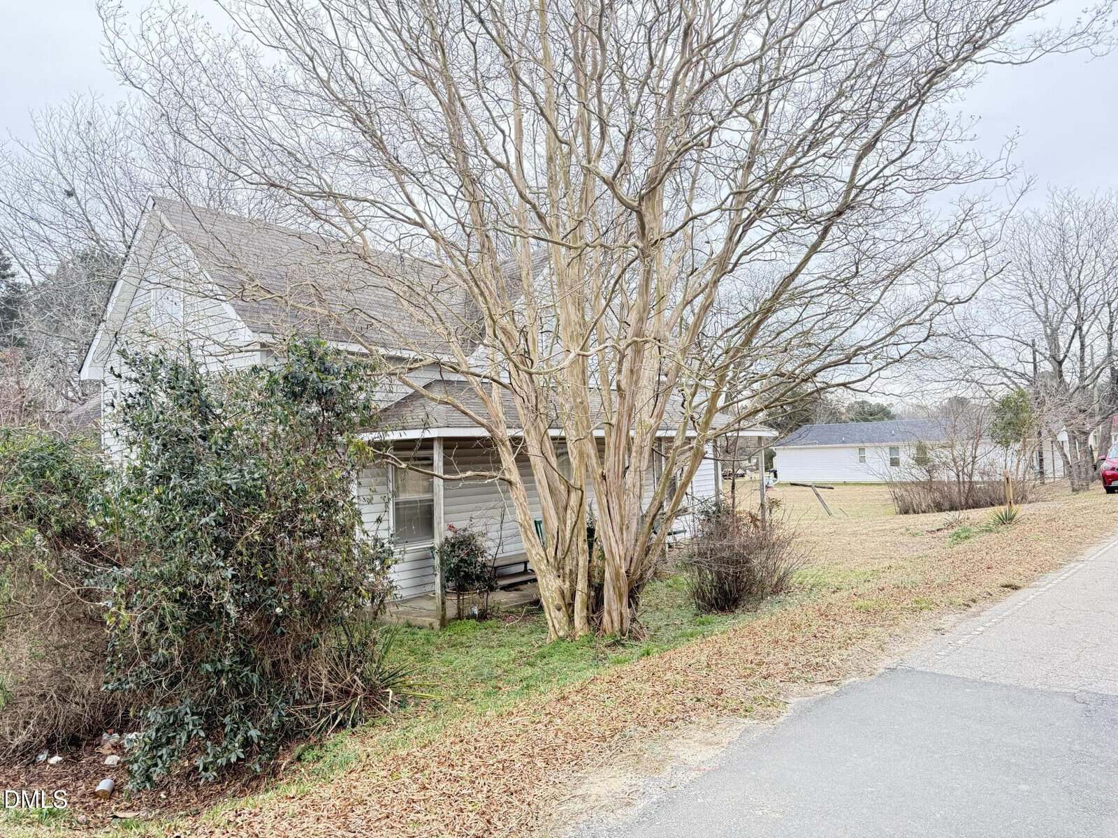 A small, weathered white house with a covered porch sits behind a leafless tree and bushes, next to a quiet street with grass and leaves scattered on the ground. Other houses and a red car are visible in the background.