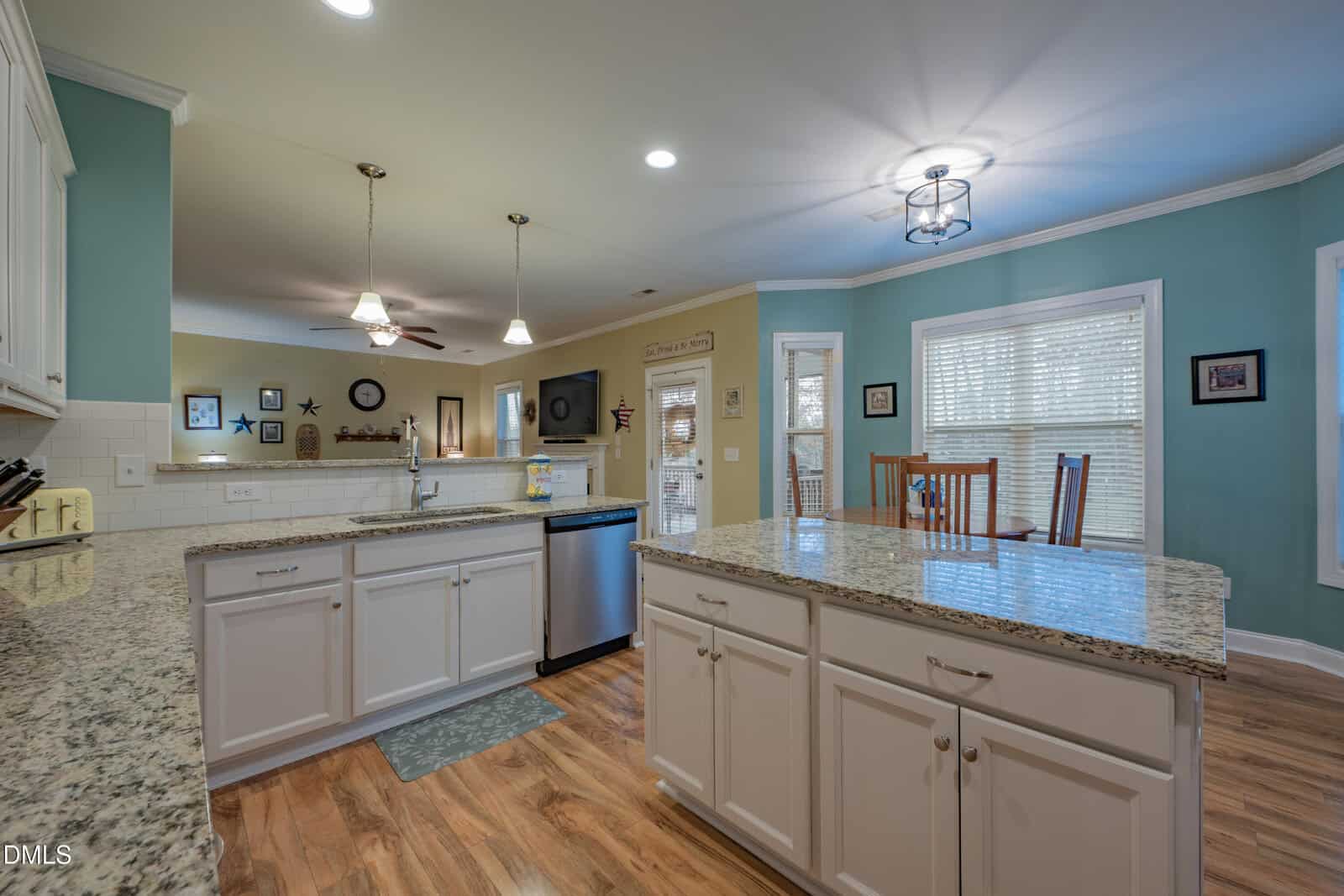 A spacious kitchen with granite countertops, white cabinets, wood floors, pendant and ceiling lights, and blue walls. There’s an island, dining table, and windows with blinds. The kitchen opens into a living area.
