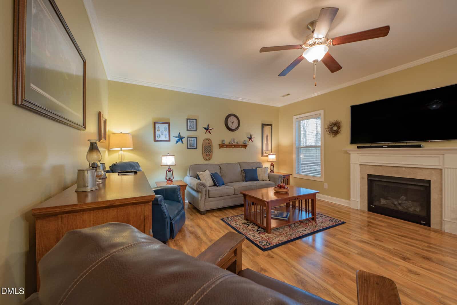 Cozy living room with a ceiling fan, two sofas, lamps, a fireplace, TV, wall decor, and wooden floor. Coffee table sits on a small rug in the center. Warm yellow walls and framed artwork complete the space.