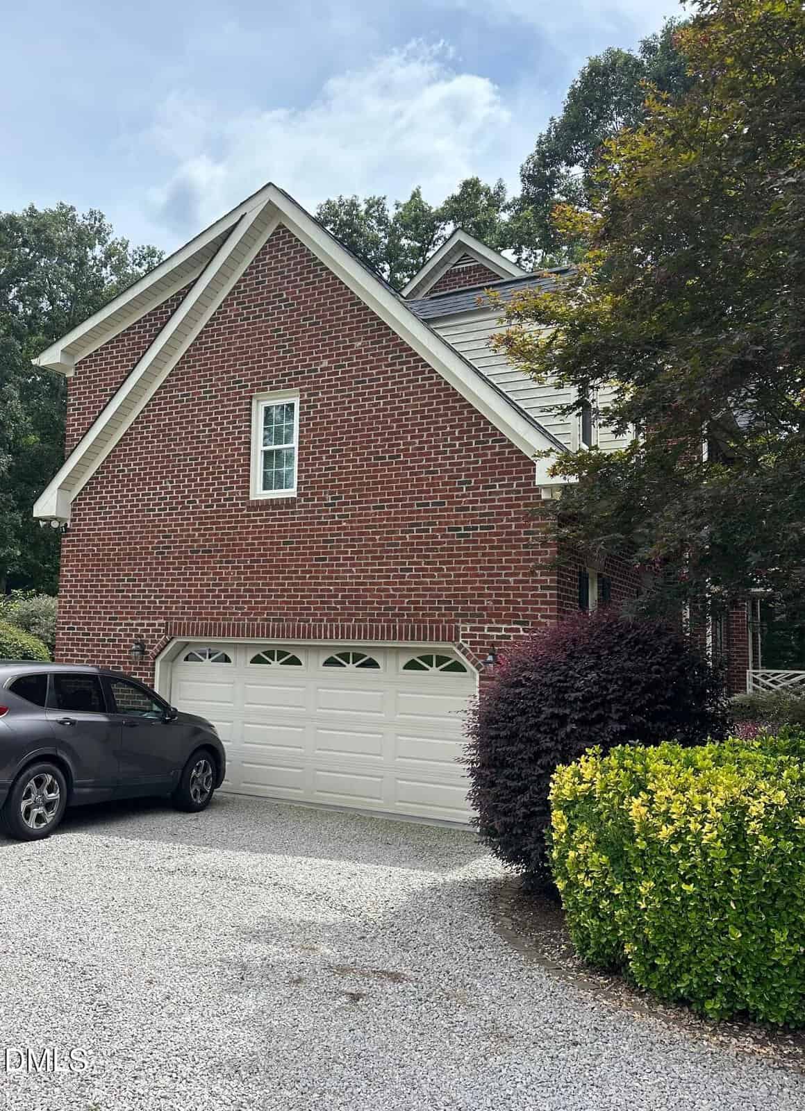A two-story red brick house with a white double garage door, a gray car parked in the gravel driveway, and bushes with green and dark red leaves beside the driveway. Trees and a partly cloudy sky are visible in the background.