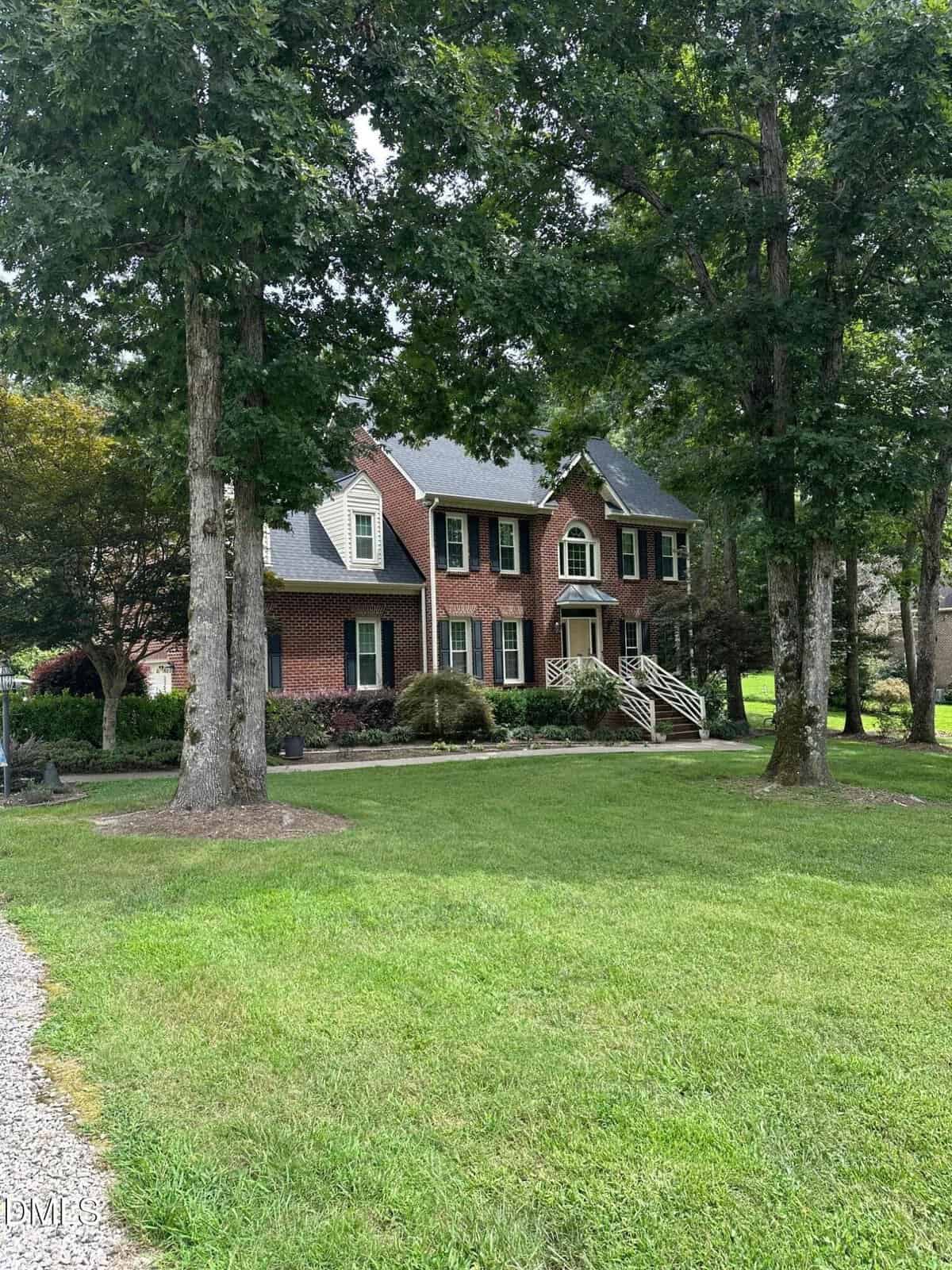 A two-story red brick house with white trim, black shutters, and a front porch with white railings sits gracefully at 5626 Marian Lane in Oxford. Mature trees and a well-kept lawn surround the house, while a gravel driveway is visible on the left.