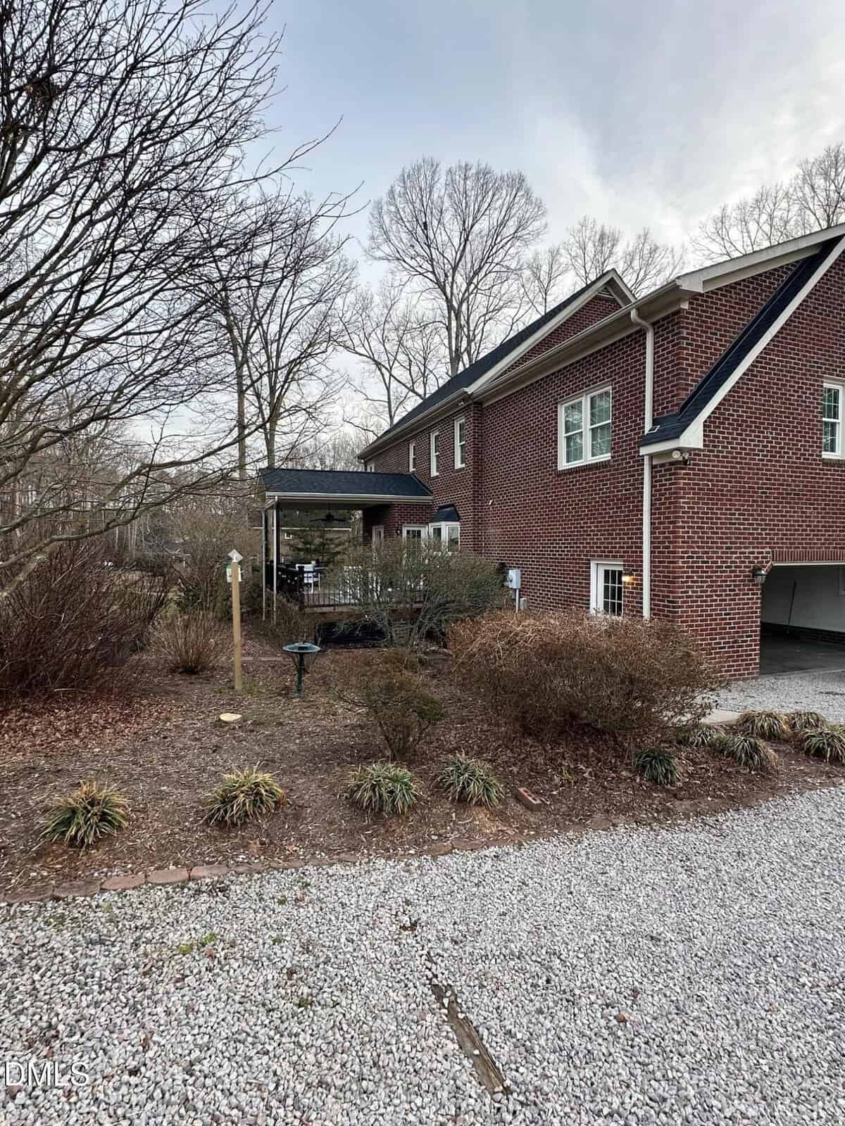 A two-story red brick house with a covered porch and white-trimmed windows sits beside a gravel driveway, with leafless trees and winter shrubs in the yard under an overcast sky.
