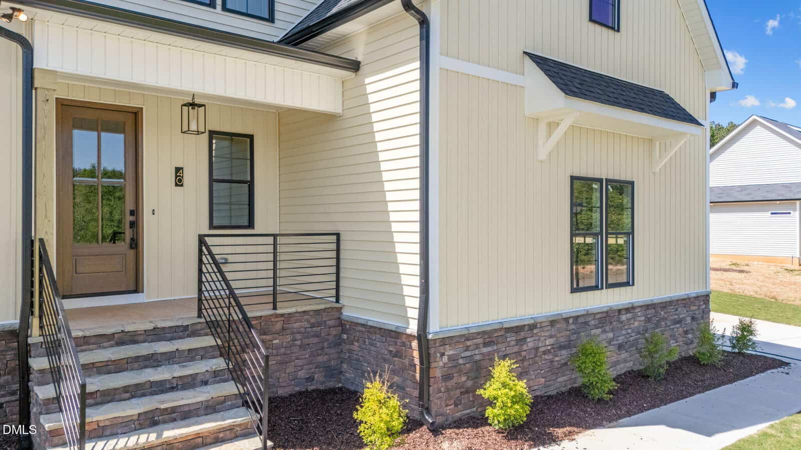 A modern house with light yellow vertical siding, stone accents at the base, a covered porch with steps, black railings, and neatly landscaped shrubs in front.