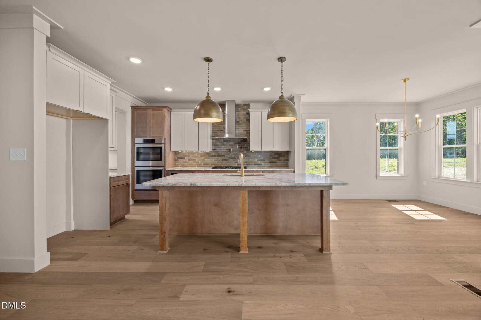 Modern kitchen with a large island, marble countertop, two pendant lights, double oven, wood floors, light cabinets, brick backsplash, and large windows letting in natural light.