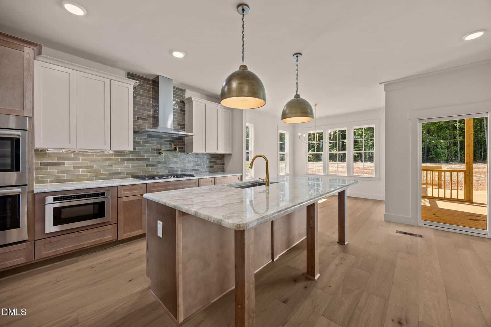 Modern kitchen with marble island, gold pendant lights, stainless steel appliances, white cabinets, tile backsplash, light wood floors, large windows, and a view of a deck and trees outside.