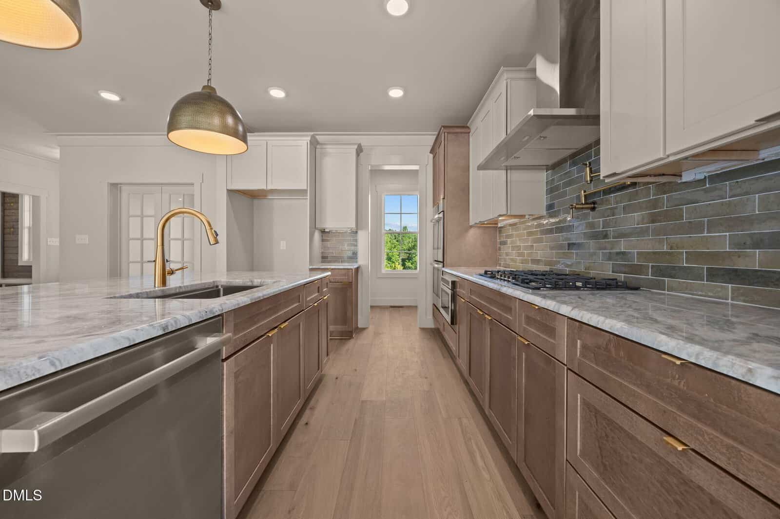 Modern kitchen with marble countertops, stainless steel appliances, brown lower cabinets, white upper cabinets, pendant lighting, and a window at the end of the room letting in natural light.