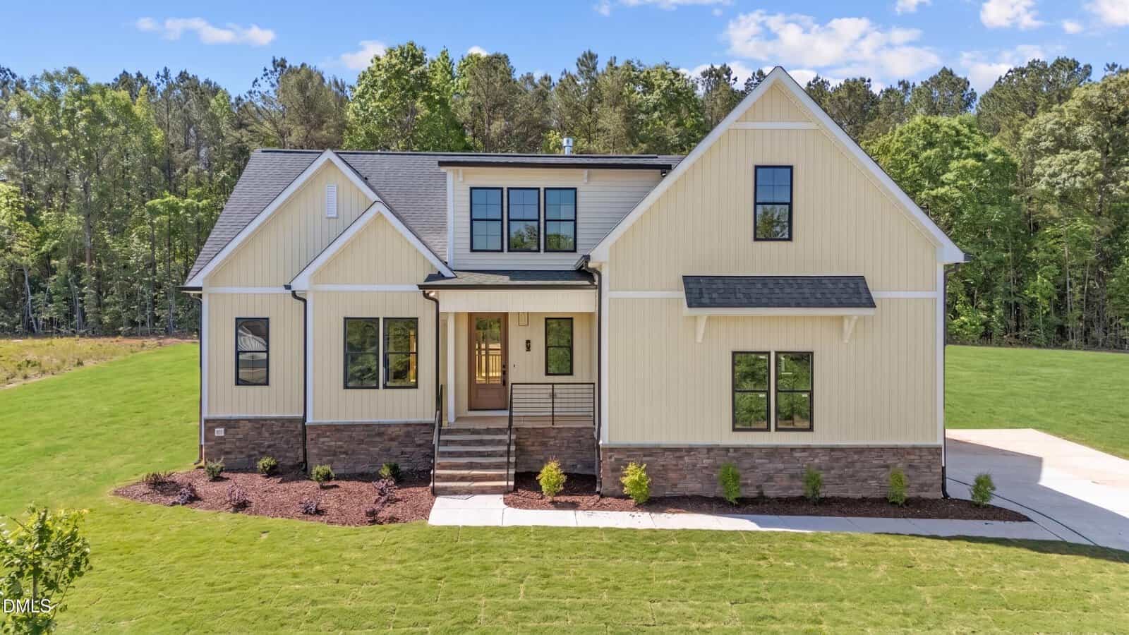 A modern, light yellow farmhouse-style home with black trim and a stone foundation, featuring large windows, a front porch with steps, and surrounded by a manicured lawn and trees in the background.