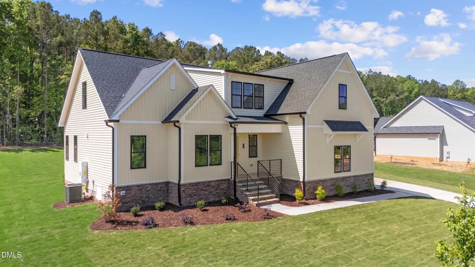 Modern beige two-story house with black trim, multiple roof peaks, and a front porch with metal railing. The home sits on a manicured lawn with young landscaping and is surrounded by trees under a blue sky.