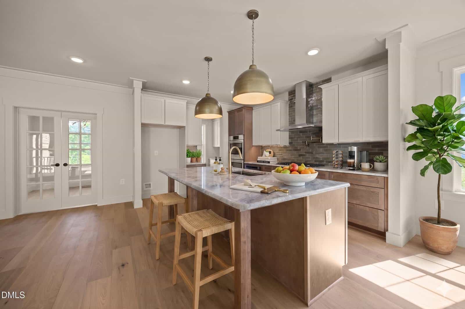 Modern kitchen with white and wood cabinets, marble island with two wooden stools, pendant lights, stainless steel appliances, fruit bowl, and a potted plant by a sunny window. French doors open to an adjacent room.