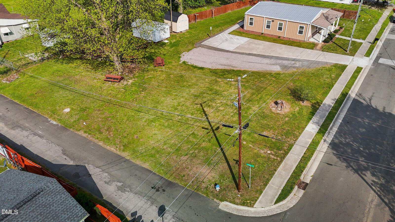 A grassy vacant lot on a street corner in Oxford, located at 216 A & B Orange Street, with a picnic table, utility poles, and power lines. Nearby are a tan building, a shed, a wooden fence, and paved roads forming an intersection.