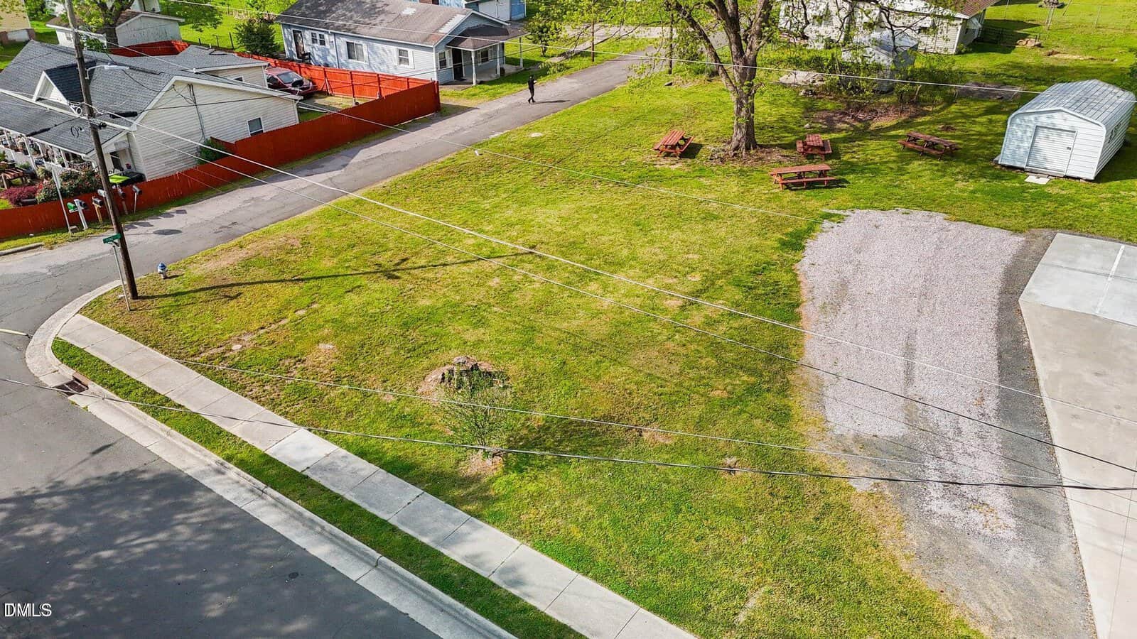 A grassy, vacant corner lot with a gravel area, picnic tables, a metal shed, overhead power lines, and sidewalks. Residential houses and a red fenced yard are visible in the background.