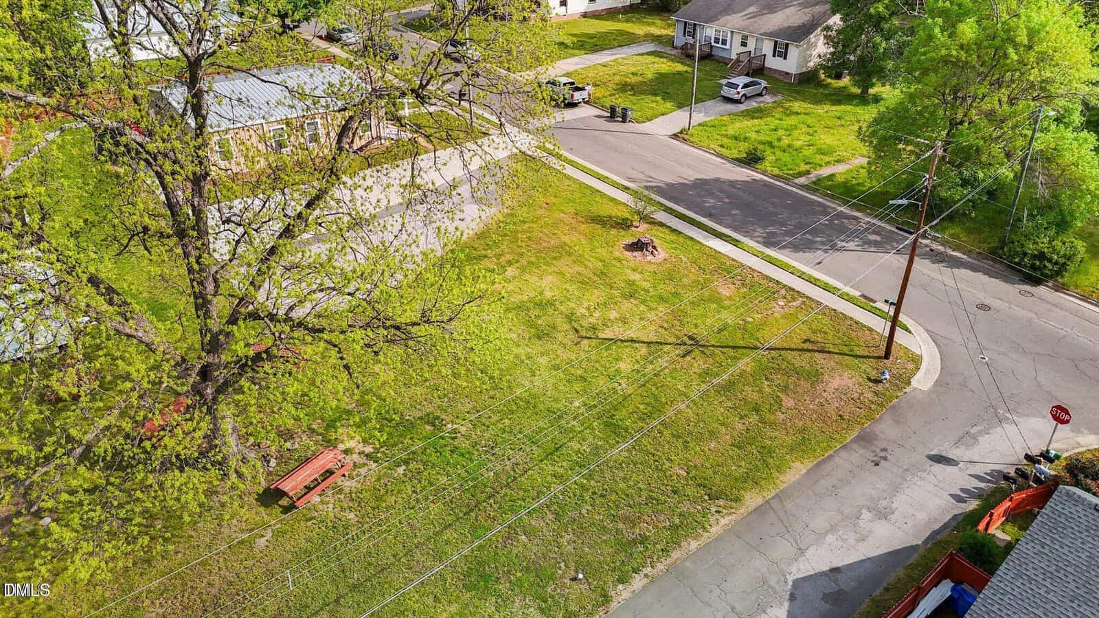 A grassy vacant lot at the corner of two residential streets, bordered by sidewalks. There is a bench near the road, overhead utility lines, a tree, several houses, and a stop sign at the intersection.