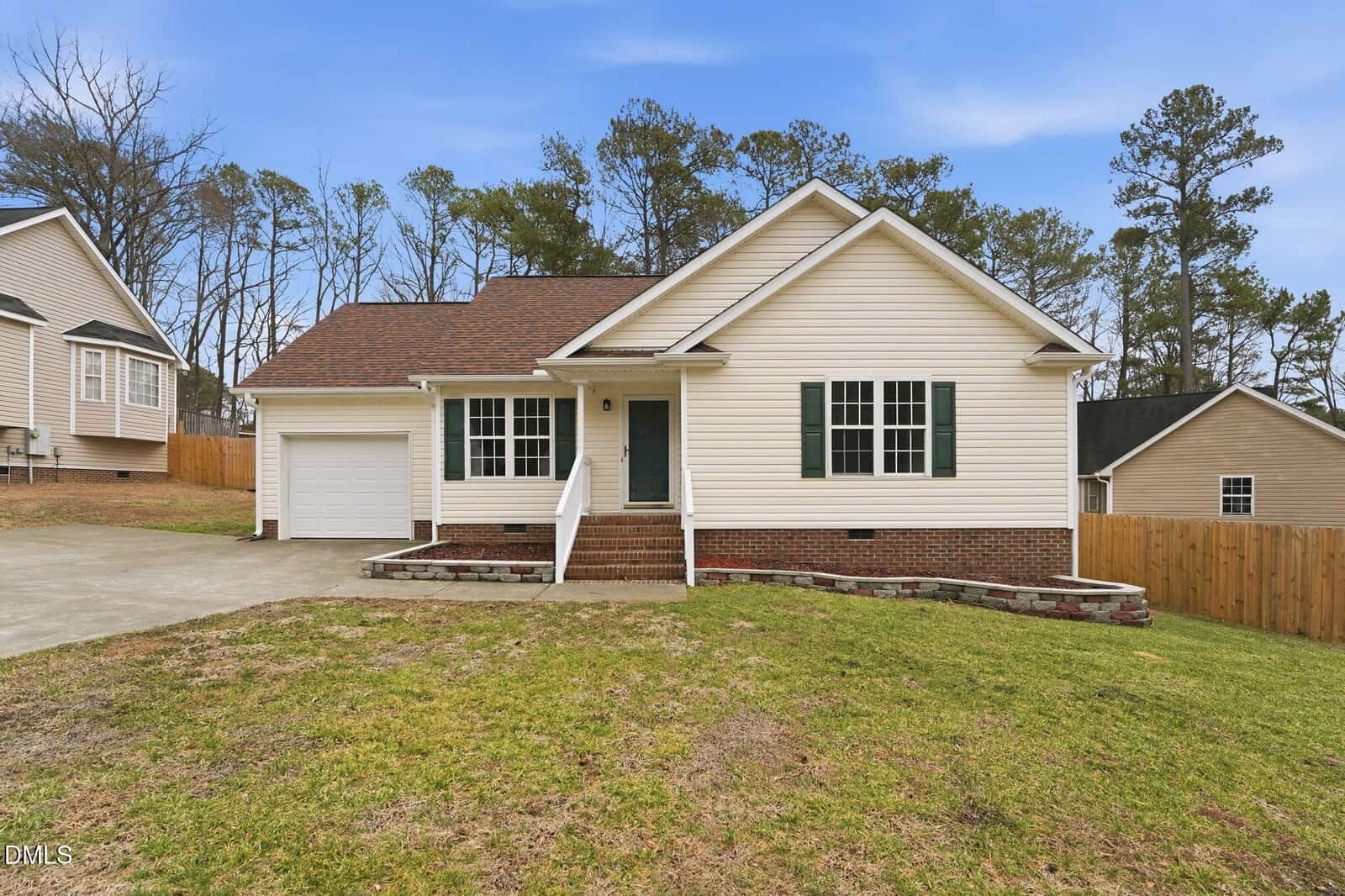 Charming single-story beige house at 104 Margaret Drive in Creedmoor, featuring a welcoming front porch, green shutters, brick foundation, attached garage, and concrete driveway. The yard has patchy grass and is enclosed by a wooden fence, with trees visible in the background.