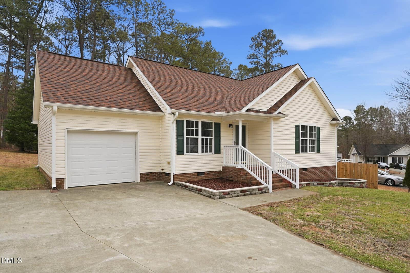 Single-story beige house with a brown shingle roof, attached one-car garage, white trim, front porch with white railing, and a concrete driveway. The lawn is partially grassed, and trees are in the background.