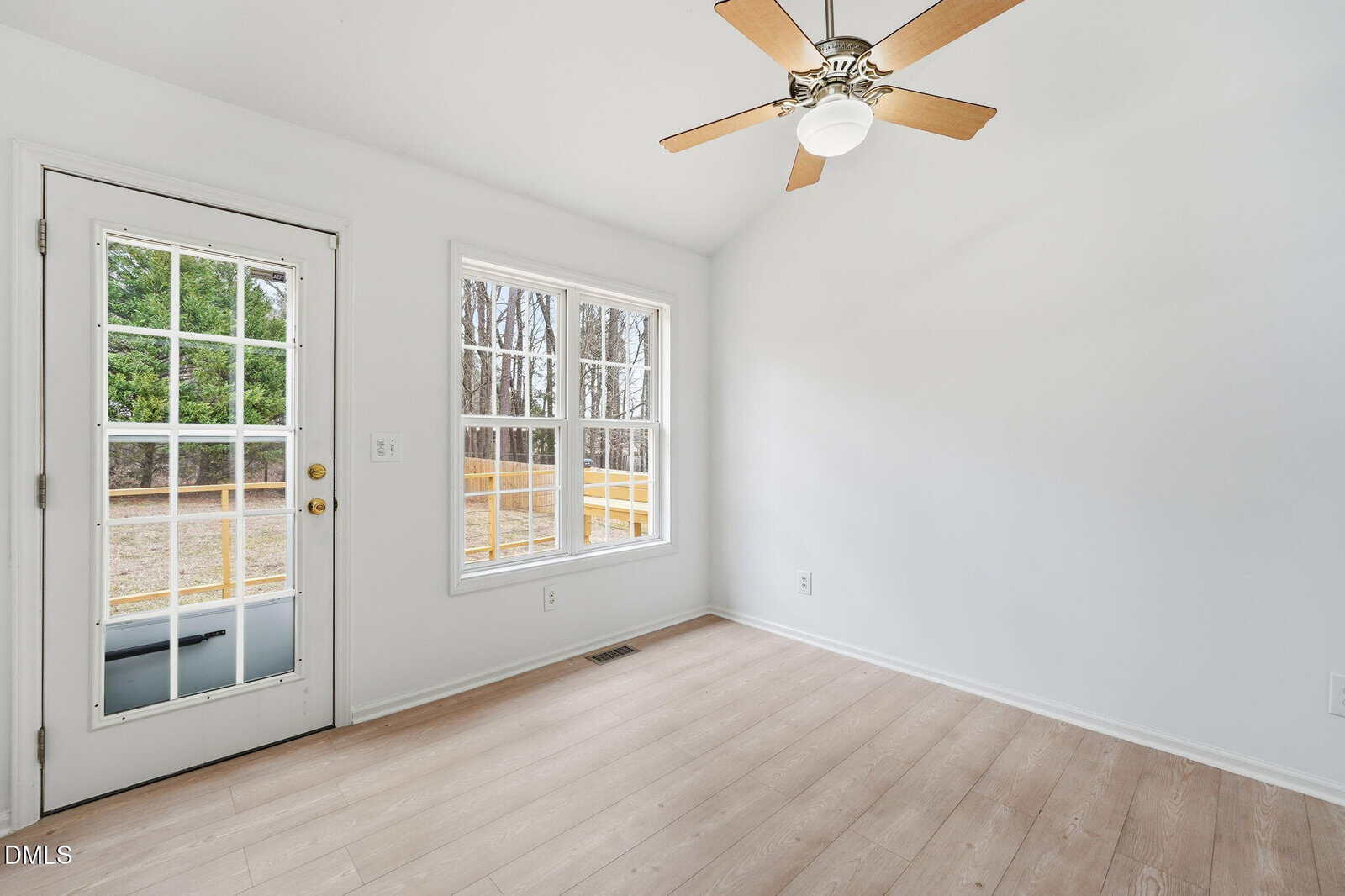 A bright, empty room with light wood floors, white walls, a ceiling fan with a light, a window, and a glass-paned door leading outside. Trees and a yellow fence are visible through the window and door.