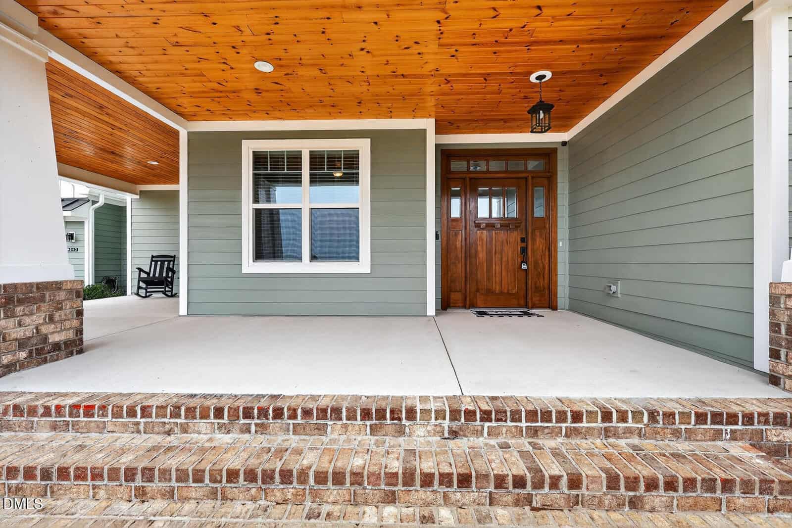 A wide, covered front porch with brick steps, light green siding, a wood-paneled ceiling, a window with white trim, and a wooden front door with glass panels. A black rocking chair is visible to the left.
