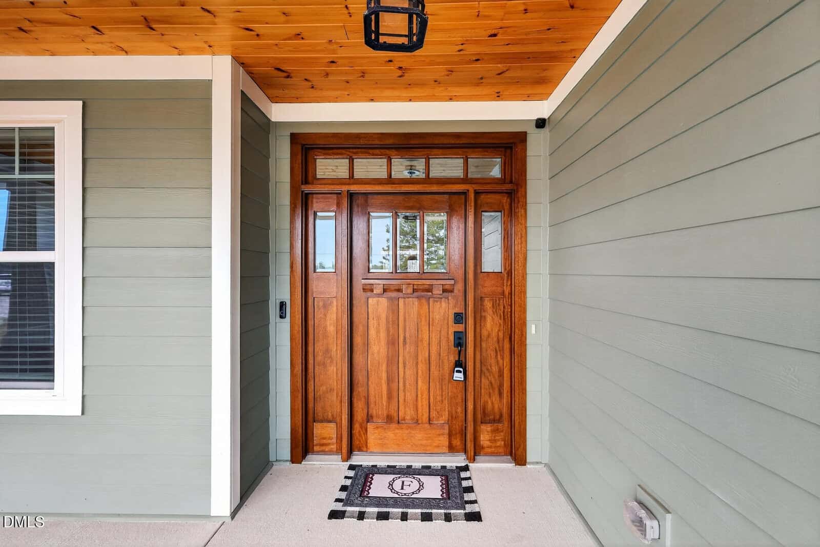 A wooden front door with glass panels and a black handle, set in a green-paneled exterior wall with white trim. A doormat with a decorative pattern is placed in front of the door. A window is visible on the left.