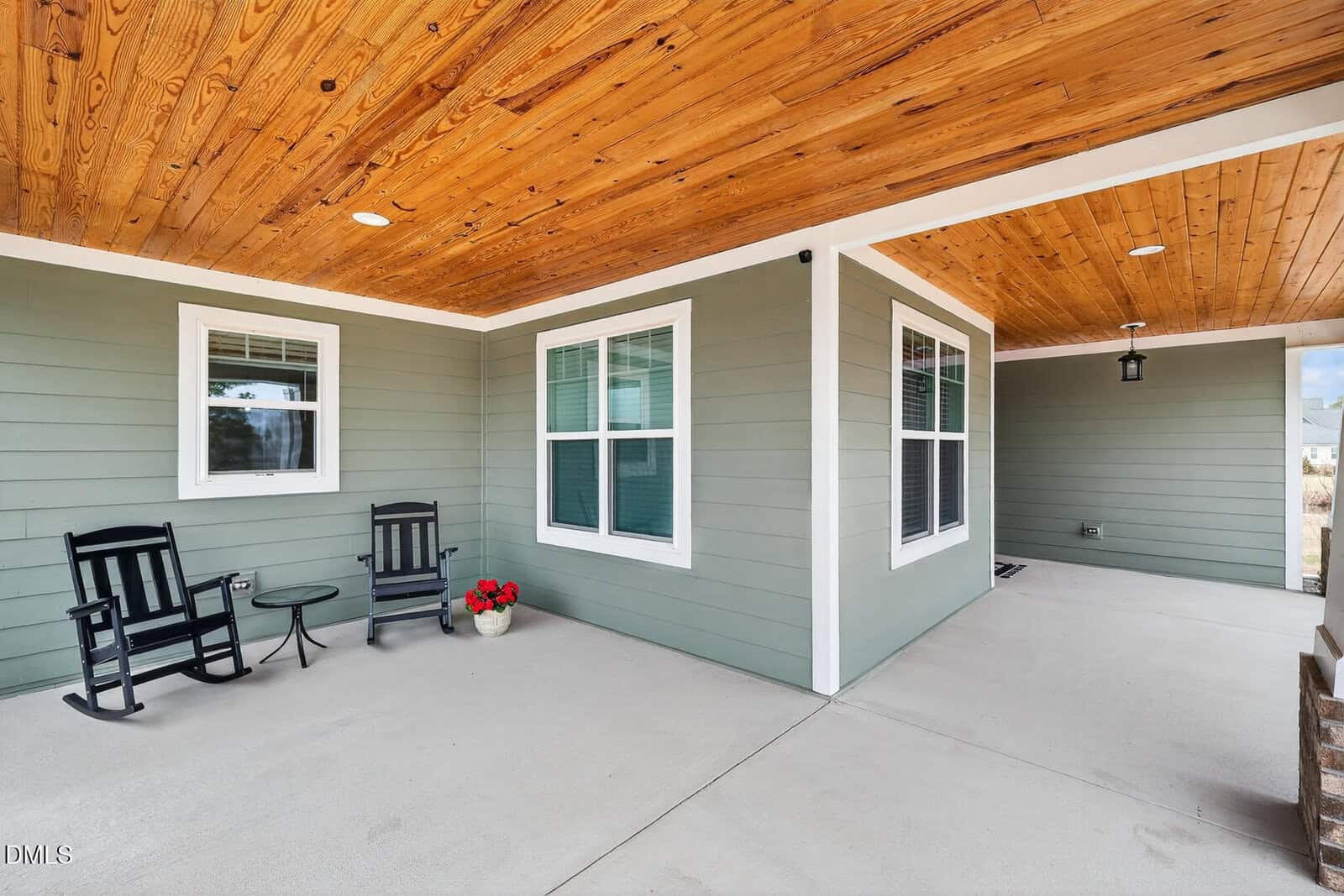 A covered porch with a wooden ceiling, light green siding, two black rocking chairs, and a pot of red flowers on a concrete floor. Multiple windows and a lantern-style light fixture are visible.