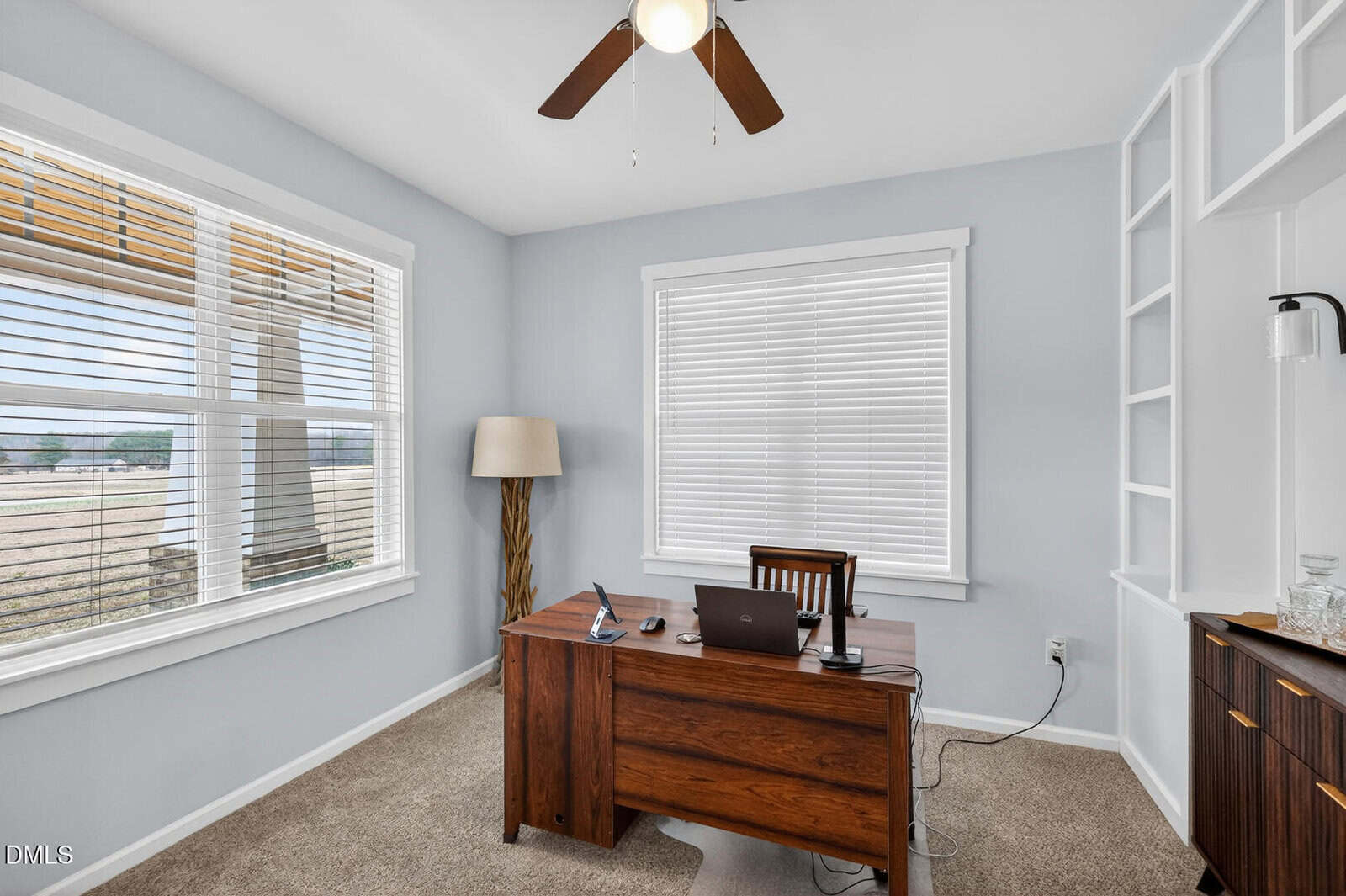A home office with a wooden desk, laptop, chair, lamp, and shelving. The room has two large windows with blinds, pale blue walls, beige carpet, and a ceiling fan with wooden blades.