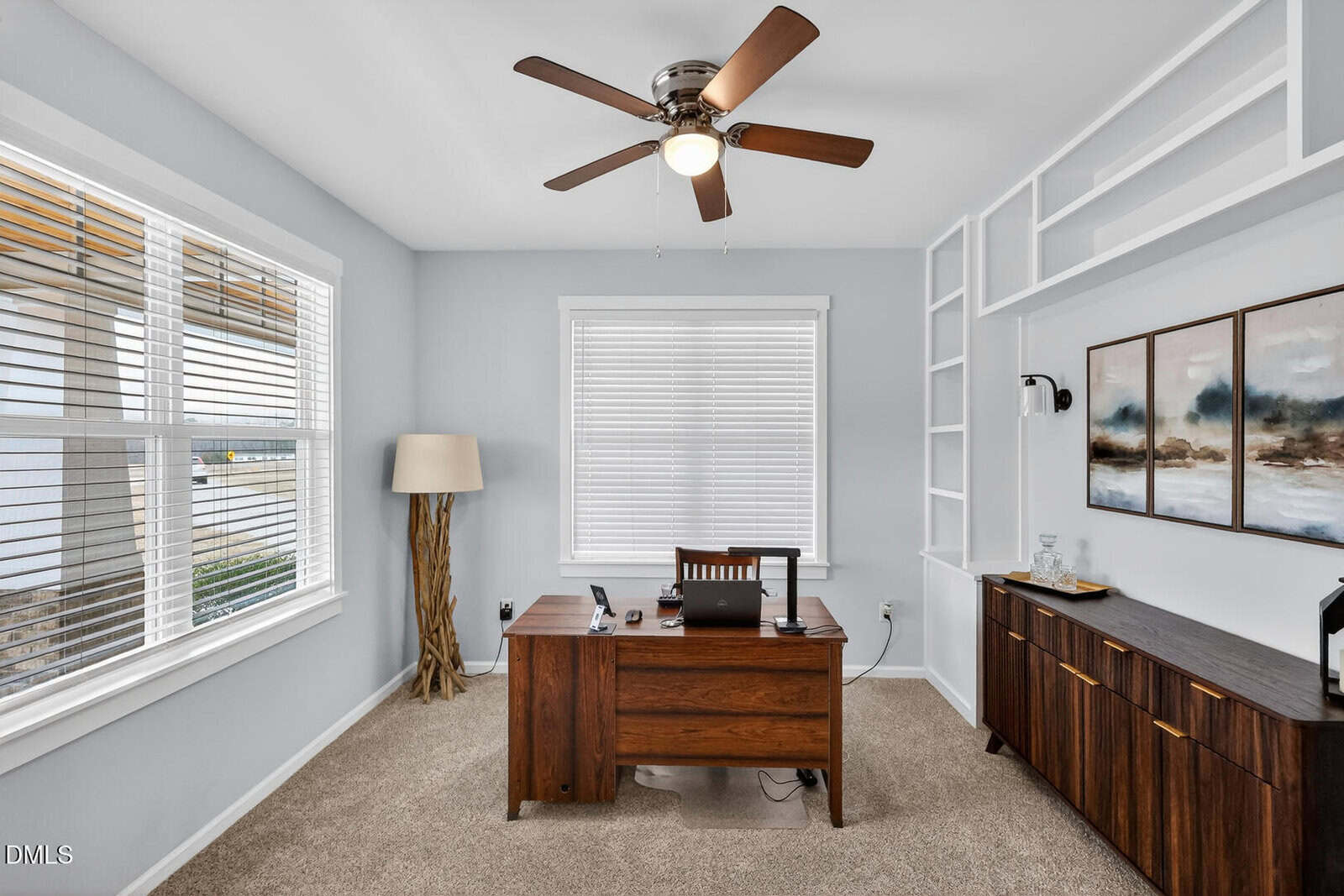 A home office with a wooden desk, laptop, and chair sits center. There is a large window with blinds, a floor lamp, a ceiling fan, a sideboard with decor, and neutral walls with built-in shelves and two paintings.
