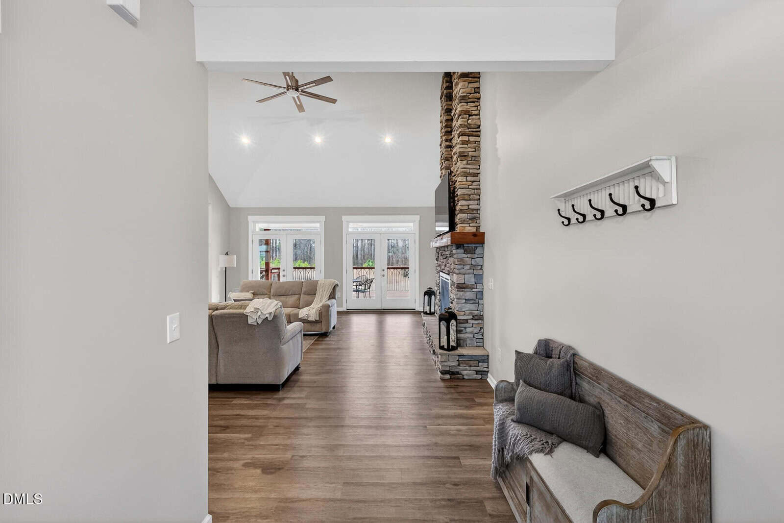 A view from an entryway into a living room with light walls, wood flooring, and a vaulted ceiling. There is a wooden bench with pillows and a coat rack on the right, and a stone fireplace in the living area ahead.