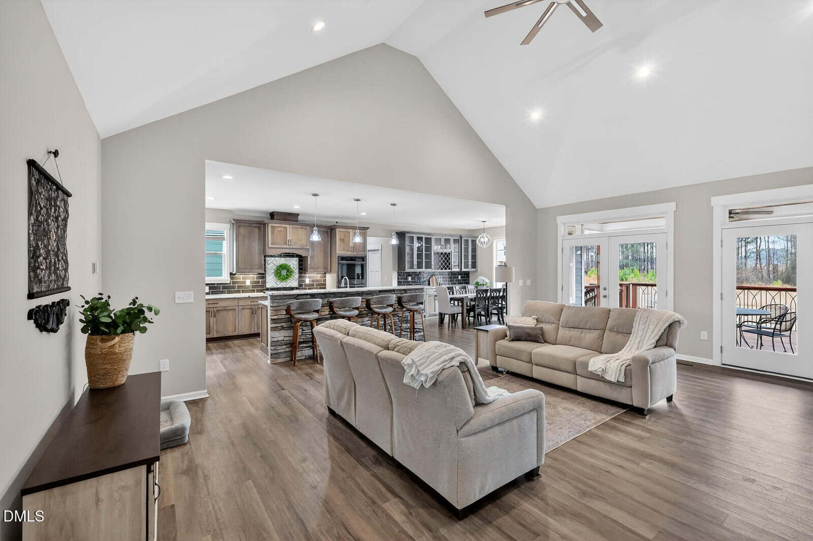 Open-concept living room with two beige sofas, wood flooring, and neutral walls. The space leads to a kitchen with island seating and a dining area. Large windows and glass doors provide natural light and outdoor views.